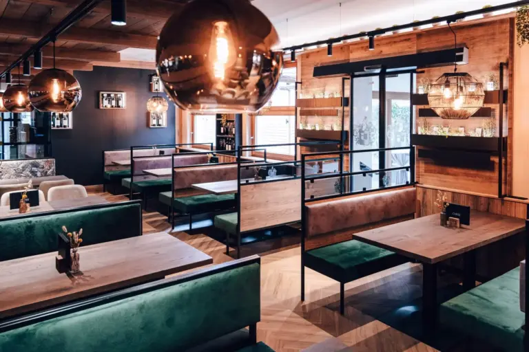 Ein modernes Restaurant mit grün gepolsterten Stühlen, Holztischen, warmer Beleuchtung, großen Hängelampen und bodentiefen Fenstern, die natürliches Licht hereinlassen. Der Raum hat eine gemütliche und einladende Atmosphäre.