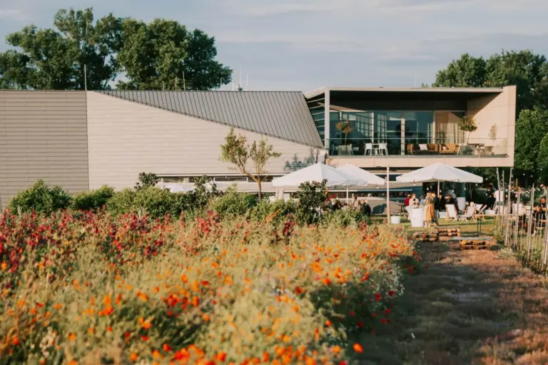Ein modernes Gebäude mit großen Fenstern steht hinter einem farbenfrohen Feld mit blühenden Blumen, weißen Sonnenschirmen und Menschen, die sich an einem sonnigen Tag draußen versammelt haben.