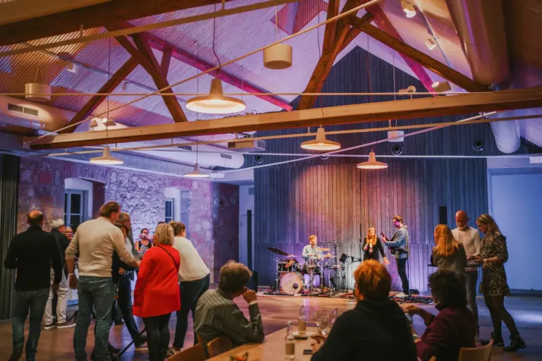 Die Leute schauen zu und tanzen, während eine Live-Band auf der Bühne in einem warm beleuchteten Raum mit Holzbalken und Steinwänden auftritt. Einige Zuschauer sitzen an Tischen, andere stehen oder tanzen in der Nähe der Bühne.