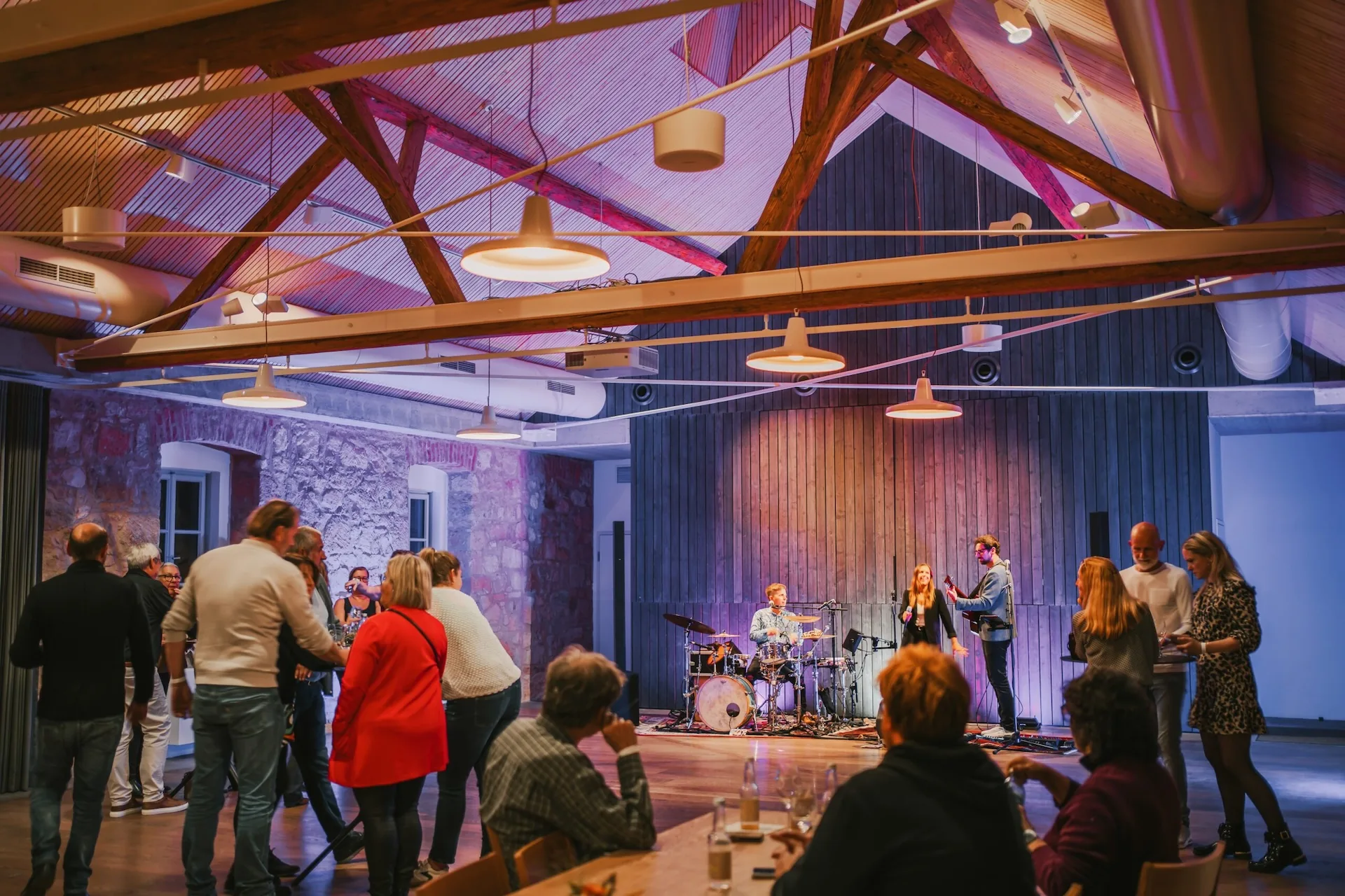 Die Leute schauen zu und tanzen, während eine Live-Band auf der Bühne in einem warm beleuchteten Raum mit Holzbalken und Steinwänden auftritt. Einige Zuschauer sitzen an Tischen, andere stehen oder tanzen in der Nähe der Bühne.