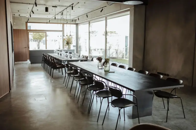 Ein modernes Esszimmer mit zwei langen, schwarzen Tischen, die parallel zueinander stehen und von schwarzen Stühlen umgeben sind. Große Fenster lassen natürliches Licht herein, und auf den Tischen stehen Vasen mit Blumen. Der Raum hat ein minimalistisches Design.