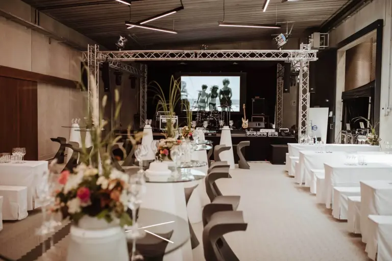 Ein moderner Veranstaltungsraum mit weißen Tischdecken, Blumenschmuck, Stehtischen und einer Bühne mit einer großen Leinwand, auf der ein Schwarz-Weiß-Bild zu sehen ist, sowie einer Licht- und Tonanlage.
