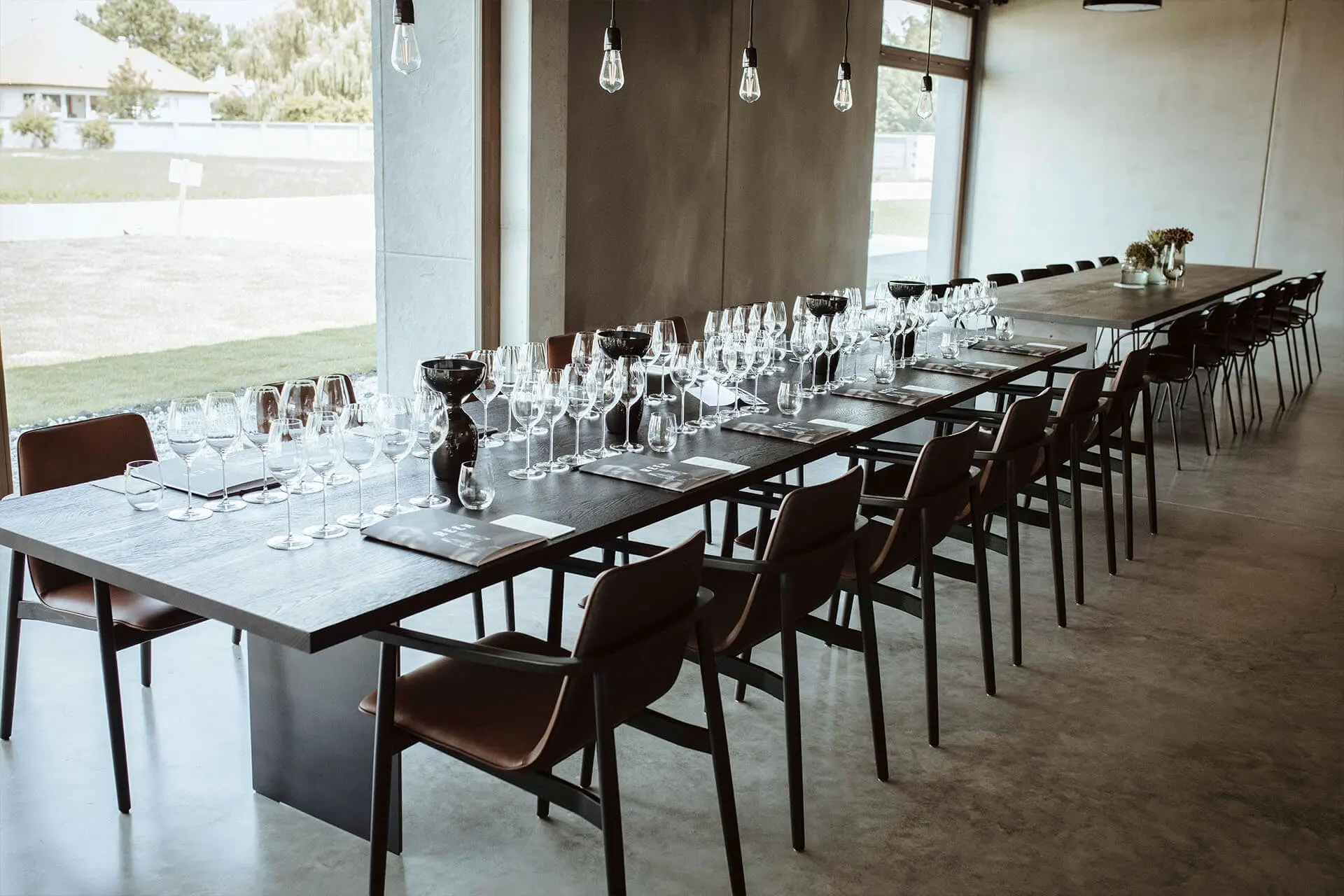 Ein langer Esstisch mit zahlreichen leeren Weingläsern und dunklen Servietten in einem modernen, minimalistischen Raum mit großen Fenstern und hängenden Glühbirnen. Die Stühle sind ordentlich an beiden Seiten des Tisches angeordnet.