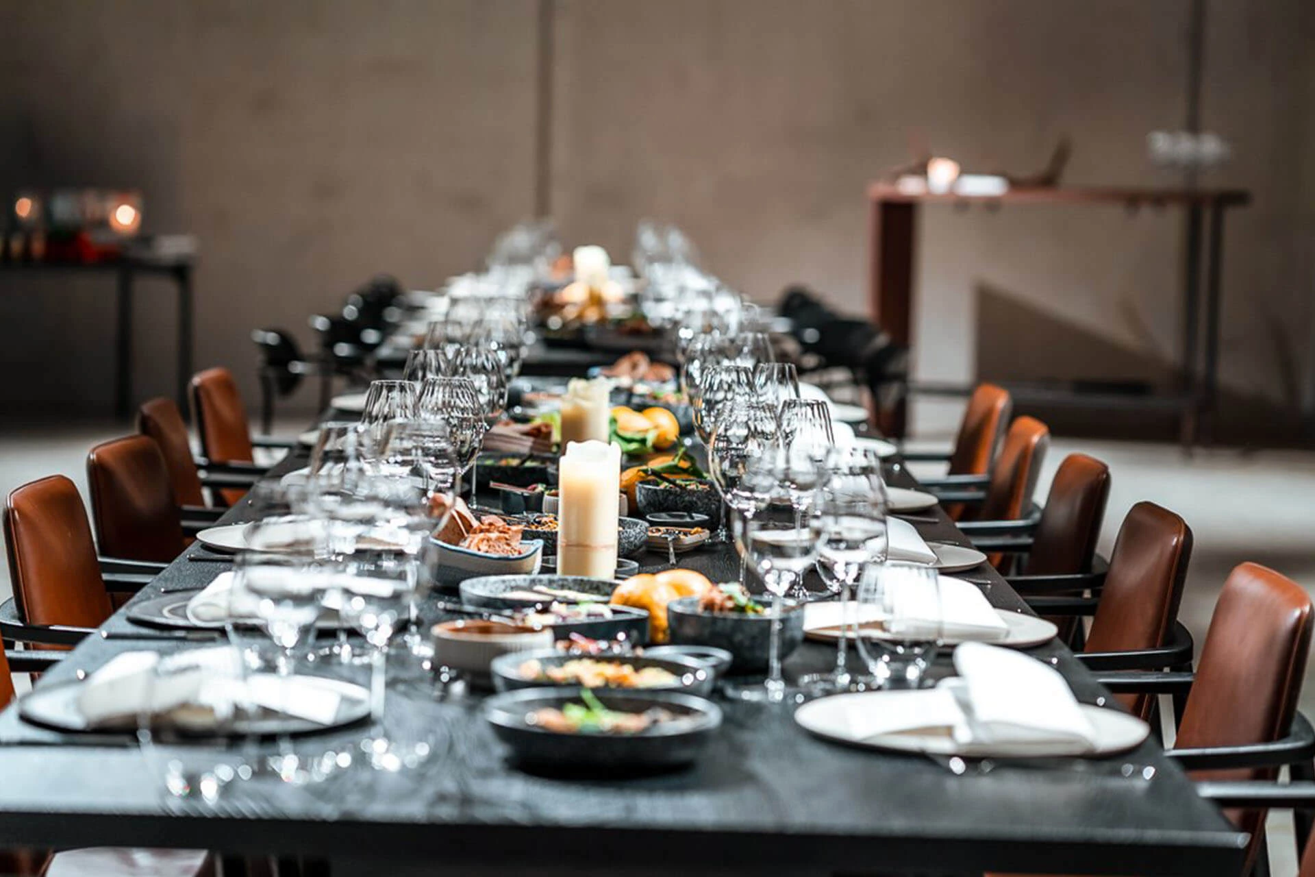 Ein langer, eleganter Esstisch, gedeckt mit Tellern, Gläsern, Servietten und verschiedenem Geschirr, mit einer brennenden Kerze in der Mitte. Auf beiden Seiten stehen braune Stühle, und der Hintergrund zeigt einen Beistelltisch in einem sanft beleuchteten Raum.