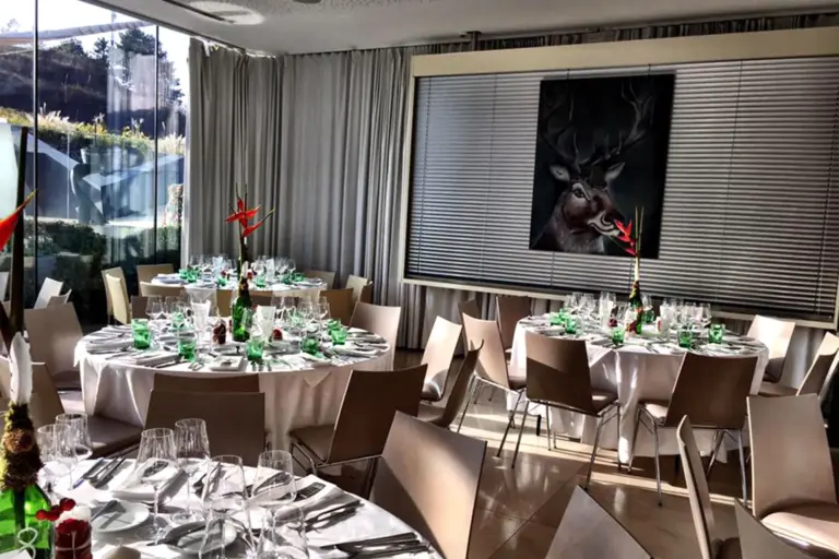 Ein moderner Speisesaal für eine Veranstaltung mit runden, weiß gedeckten Tischen, ordentlich arrangierten Stühlen, Gläsern, grünen Flaschen, roten Blumensträußen, großen Fenstern und einem Hirschgemälde an der Wand.