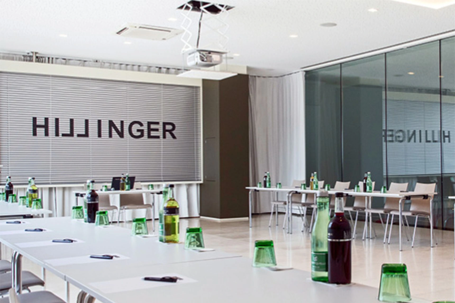 Ein moderner Konferenzraum mit langen Tischen, grünen Glastassen, Flaschen und Notizblöcken auf jedem Tisch. Der Raum hat Glaswände und Jalousien mit dem Schriftzug HILLINGER auf der Rückseite.