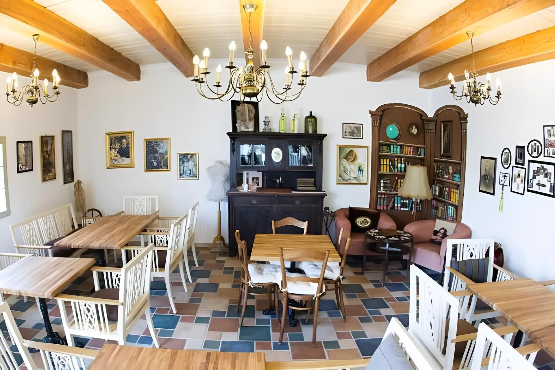 Ein gemütlicher, gut beleuchteter Raum mit Holzbalken, Kronleuchtern, weißen Stühlen und Holztischen. Ein dunkler Schrank, ein Bücherregal, ein Sofa, ein Sessel und gerahmte Kunst an den Wänden schaffen eine gemütliche, eklektische Atmosphäre.