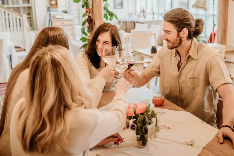 Vier Personen sitzen in einem gemütlichen, hellen Raum um einen Tisch herum, lächeln und stoßen mit einem Glas an. Der Tisch ist mit Blumen und Kerzen geschmückt, was eine warme, fröhliche Atmosphäre schafft.