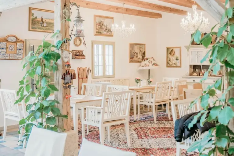 Ein gemütliches Esszimmer im Vintage-Stil mit weißen Holztischen und -stühlen, Kronleuchtern, gerahmten Kunstwerken an weißen Wänden, Zimmerpflanzen und einem gemusterten Teppich auf dem Boden. Durch ein Fenster fällt natürliches Licht ein.