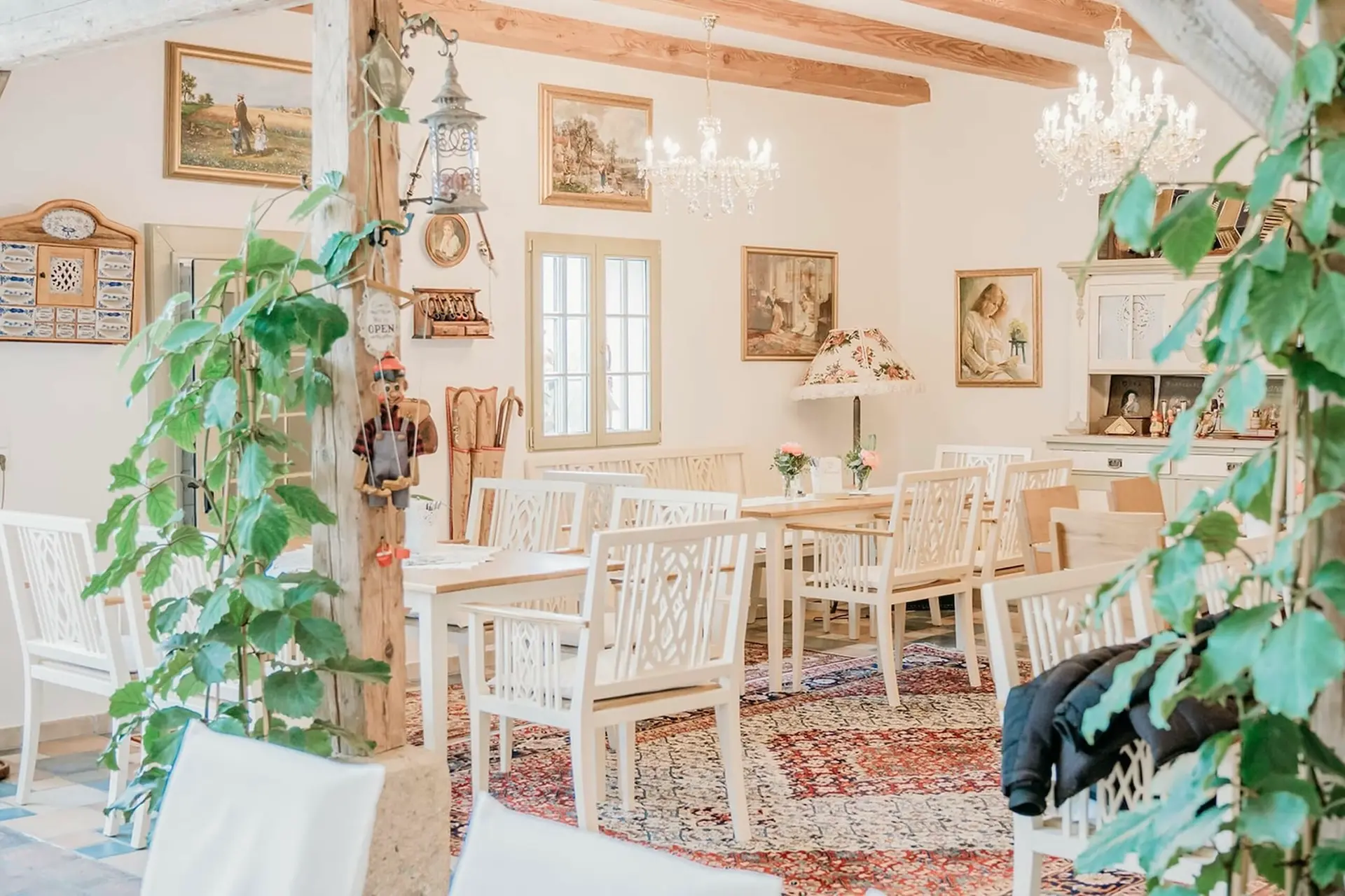 Ein gemütliches Esszimmer im Vintage-Stil mit weißen Holztischen und -stühlen, Kronleuchtern, gerahmten Kunstwerken an weißen Wänden, Zimmerpflanzen und einem gemusterten Teppich auf dem Boden. Durch ein Fenster fällt natürliches Licht ein.