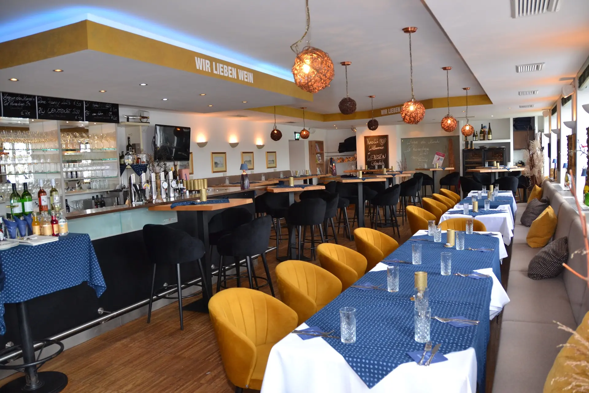 Modernes Restaurantinterieur mit gelben Stühlen, blauen Tischdecken, einem Barbereich und Pendelleuchten. An der Wand steht der deutsche Schriftzug „WIR LIEBEN WEIN“. Die Tische sind mit Gläsern und Besteck gedeckt, was eine gemütliche, einladende Atmosphäre schafft.