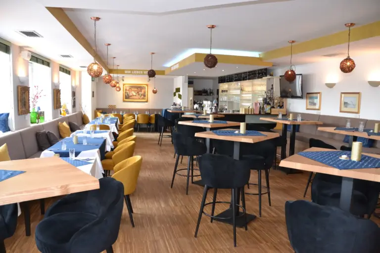 Modernes Restaurantinterieur mit Holzböden, blauen und gelben Stühlen, Tischen mit blauen Tischsets, hängenden Pendelleuchten, einem Barbereich im hinteren Bereich und Wandbildern als Dekoration.