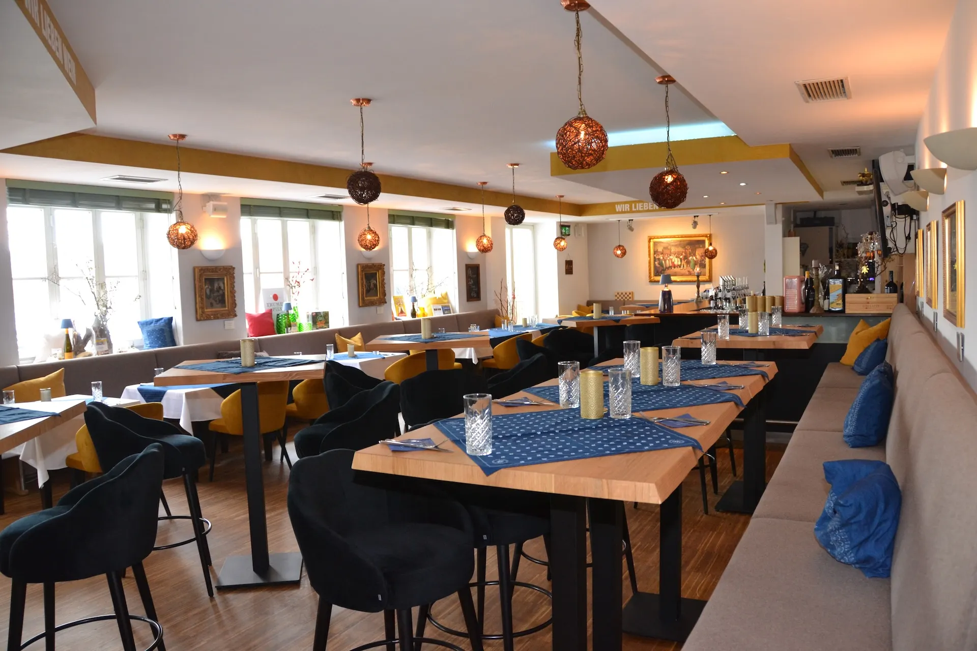 Modernes Restaurantinterieur mit Holzböden, Tischen mit blauen Tischdecken, gepolsterten Stühlen, Hängeleuchten und dekorativen Flaschen auf den Fensterbänken. Der Raum ist hell und wird durch große Fenster mit Tageslicht durchflutet.