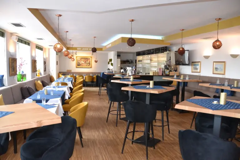 Weinstein Krems Modernes Restaurantinterieur mit Holzböden, dunklen Stühlen, eckigen Tischen, die mit blauen Tischsets und weißen Tischdecken gedeckt sind, Hängeleuchten, Wandkunst und einem Barbereich im Hintergrund.