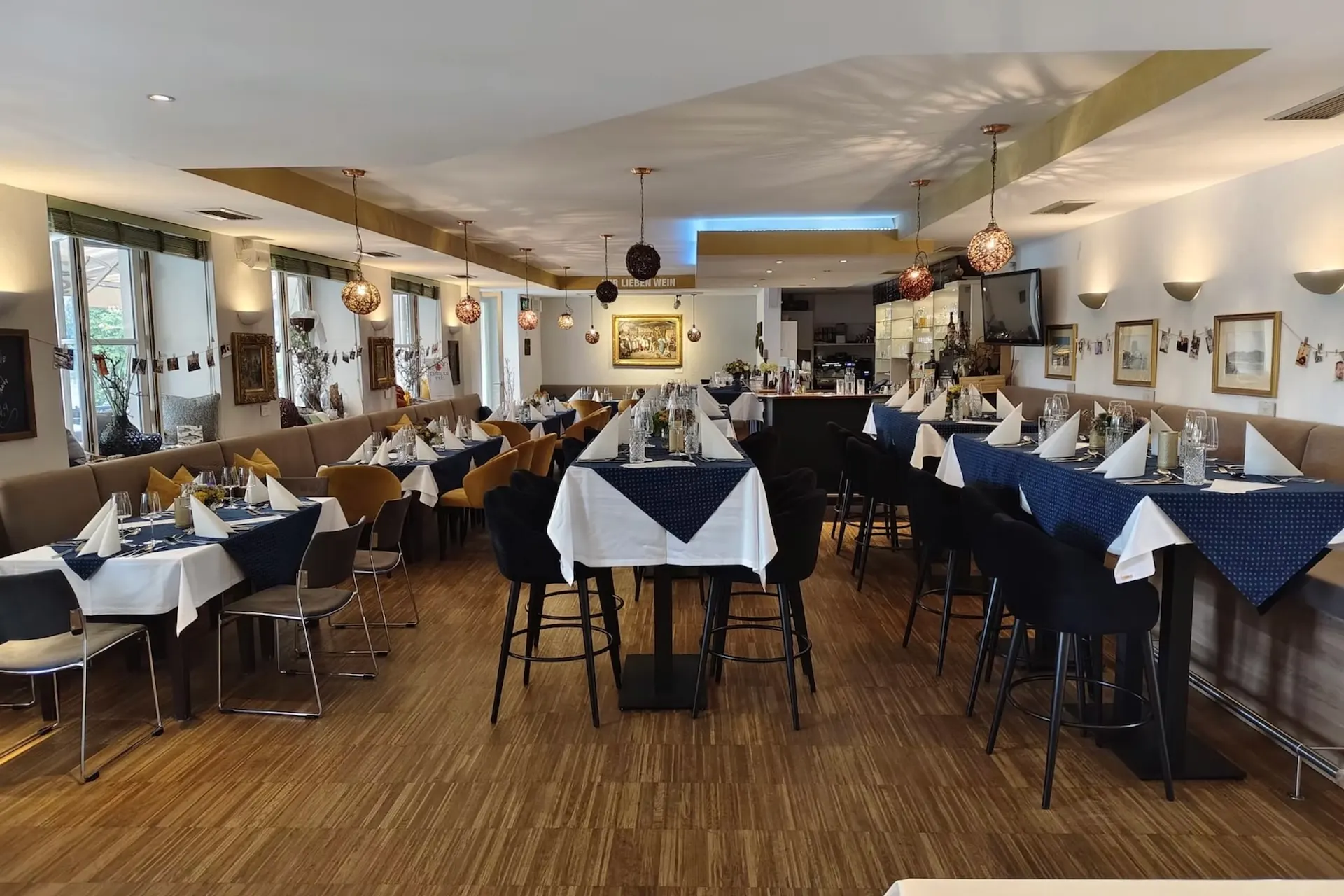 Speisesaal im WEINSTEIN KREMS mit Reihen von Tischen, die mit weißen Tischdecken und blau gemusterten Tischläufern gedeckt sind, mit Gedecken bestückt und von schwarzen Hockern flankiert.