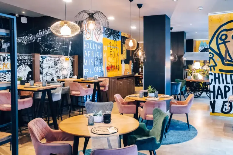 Modernes Café mit farbenfroher Wandkunst, runden Holztischen, Plüschsesseln in Rosa, Grün und Grau, Hängeleuchten und dekorativen Pflanzen, die eine lebendige und gemütliche Atmosphäre schaffen.