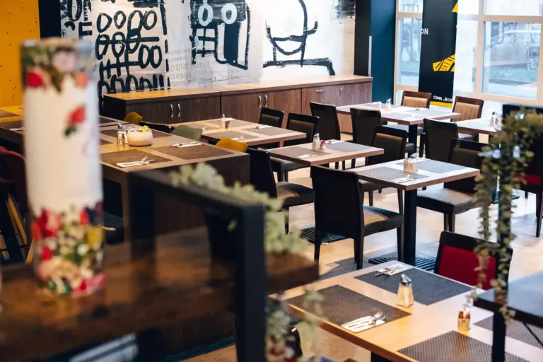 Ein moderner Restaurantbereich mit Holztischen und -stühlen, Gedecken auf jedem Tisch und abstrakten Schwarz-Weiß-Kunstwerken an der Wand im Hintergrund. Der Raum ist durch große Fenster gut mit natürlichem Licht beleuchtet.