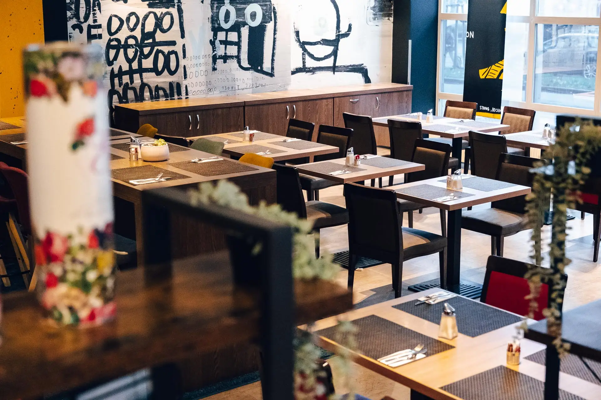 Ein moderner Restaurantbereich mit Holztischen und -stühlen, Gedecken auf jedem Tisch und abstrakten Schwarz-Weiß-Kunstwerken an der Wand im Hintergrund. Der Raum ist durch große Fenster gut mit natürlichem Licht beleuchtet.