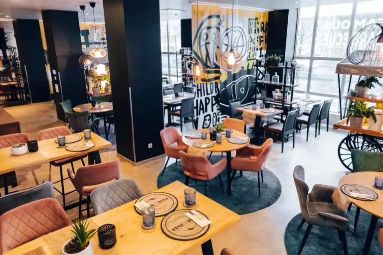 Modernes Restaurantinterieur mit Holztischen, bunten gepolsterten Stühlen, stilvoller Beleuchtung und dekorativer Wandkunst. Auf den Tischen sind Gedecke angeordnet, die eine einladende Atmosphäre zum Essen schaffen. Große Fenster lassen viel natürliches Licht herein.