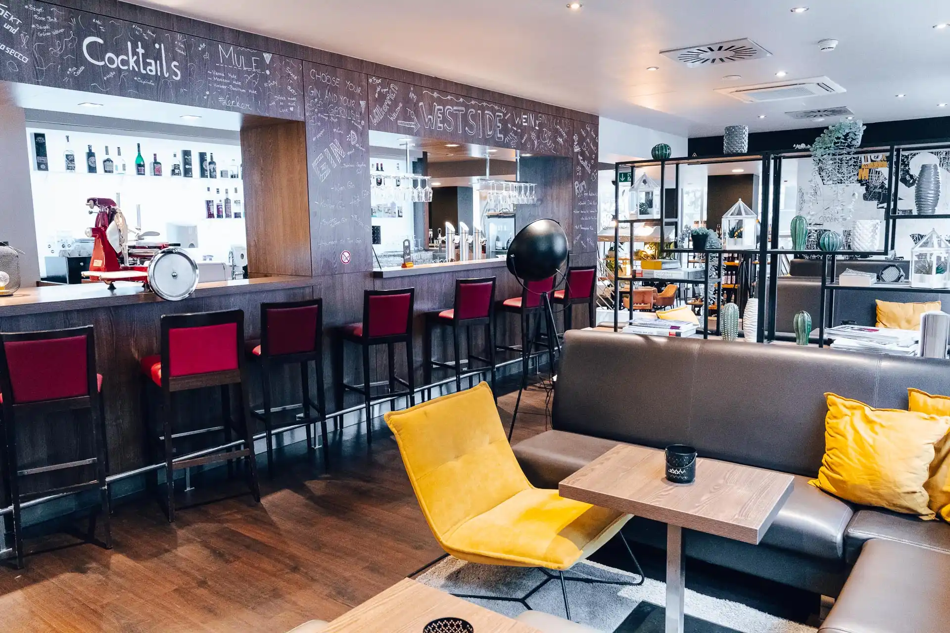 Moderne Lounge-Bar mit Holzböden, hohen Tischen und roten Stühlen an der Bar, einem gelben Sessel, grauen Sofas mit gelben Kissen und Kreidetafeln mit handgeschriebenen Nachrichten und Getränkenamen.