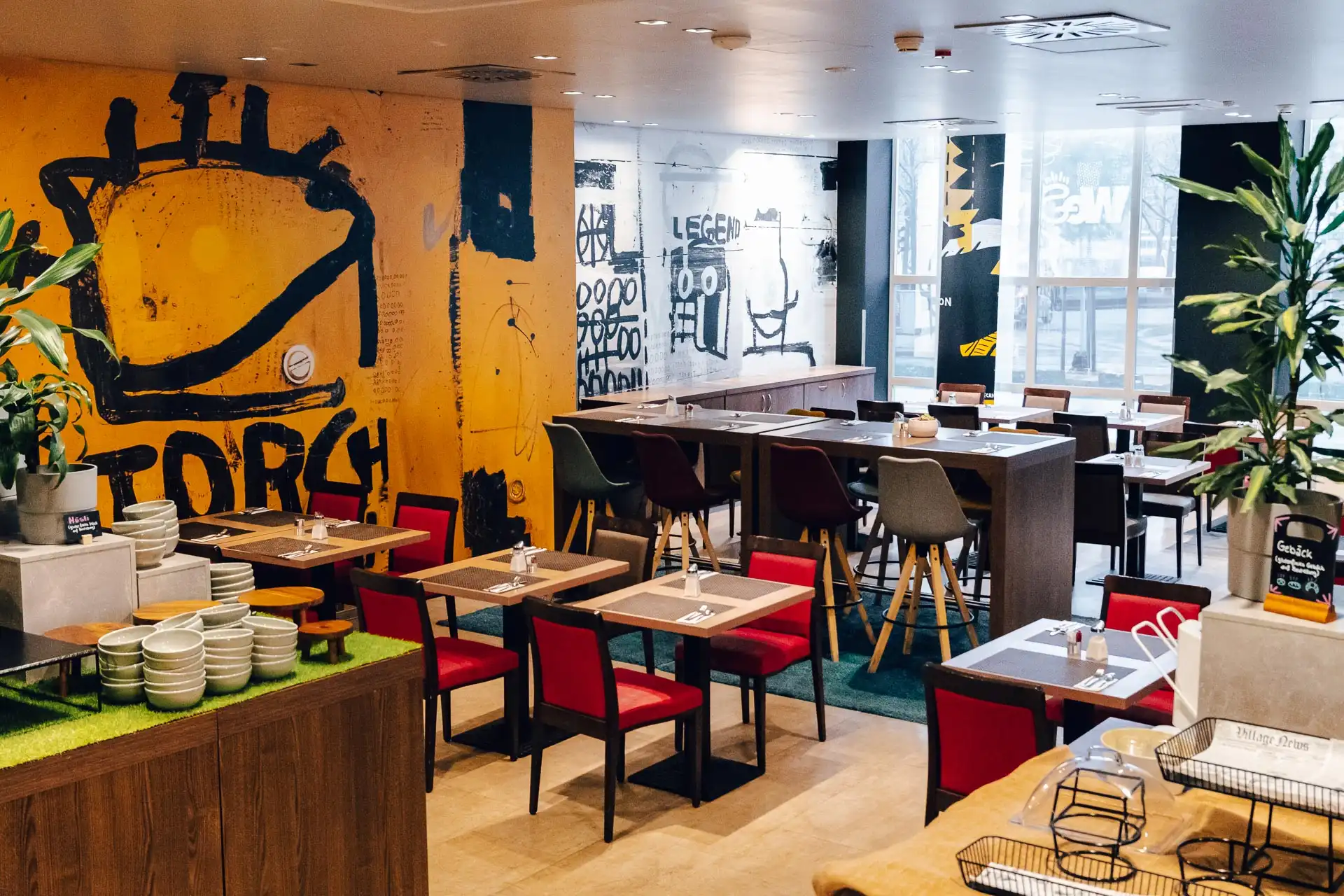 Die moderne Einrichtung des Restaurants mit Holztischen, roten und grauen Stühlen, leuchtend gelber und schwarzer Wandkunst, großen Fenstern und Grünpflanzen schafft eine lebendige und einladende Atmosphäre.
