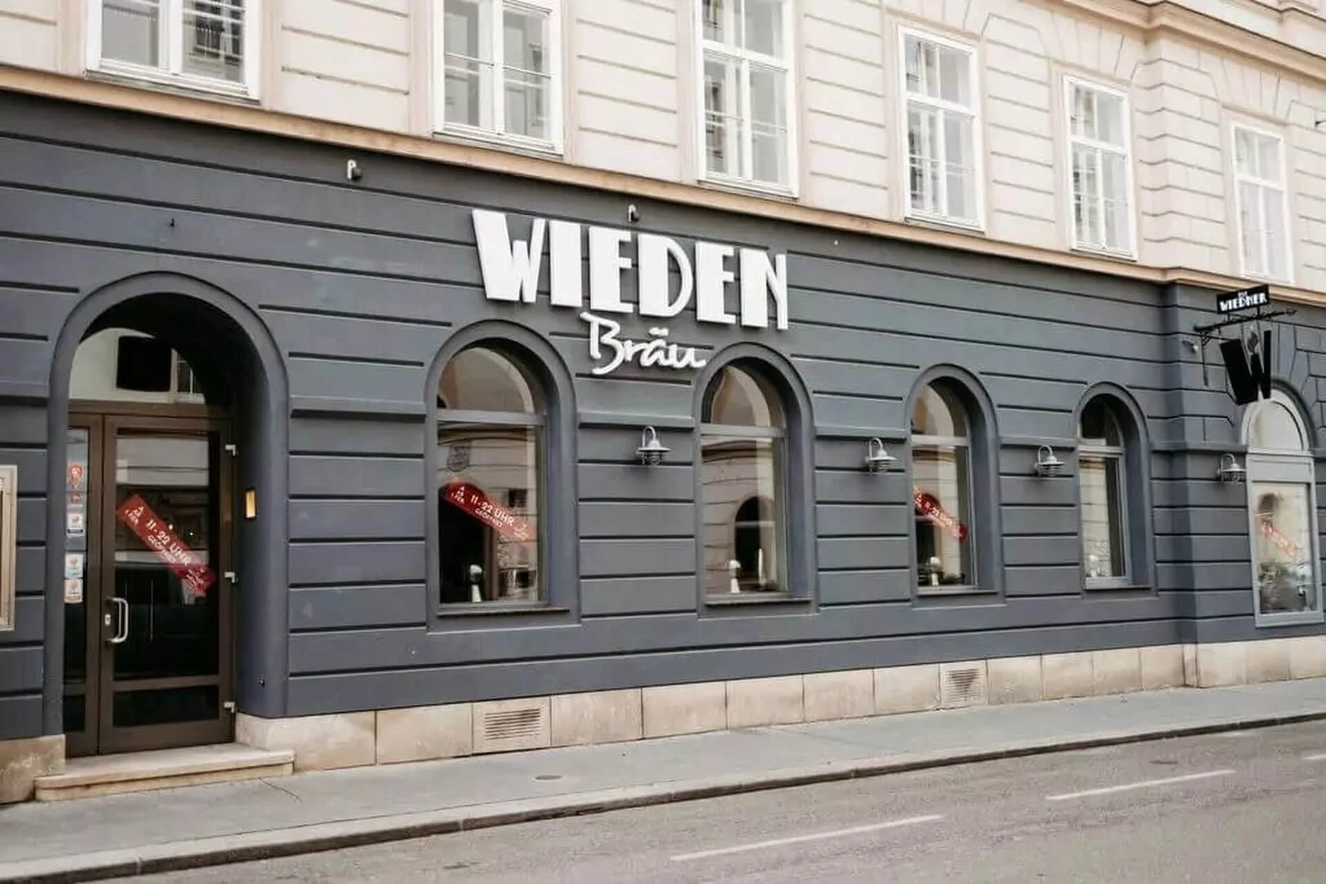 Straßenansicht eines Gebäudes mit grauen Wänden und gewölbten Fenstern, auf dem ein großes Schild mit der Aufschrift "Wieden Bräu" angebracht ist. Draußen ist keine sichtbare Aktivität zu sehen, und die Straße ist leer.