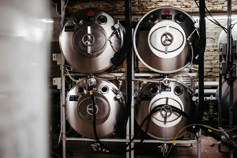 Vier große Bierfässer aus rostfreiem Stahl sind in einer Brauerei gestapelt und mit Schläuchen und Ventilen verbunden. Im Hintergrund sind eine freiliegende Ziegelwand und andere Brauereianlagen zu sehen.