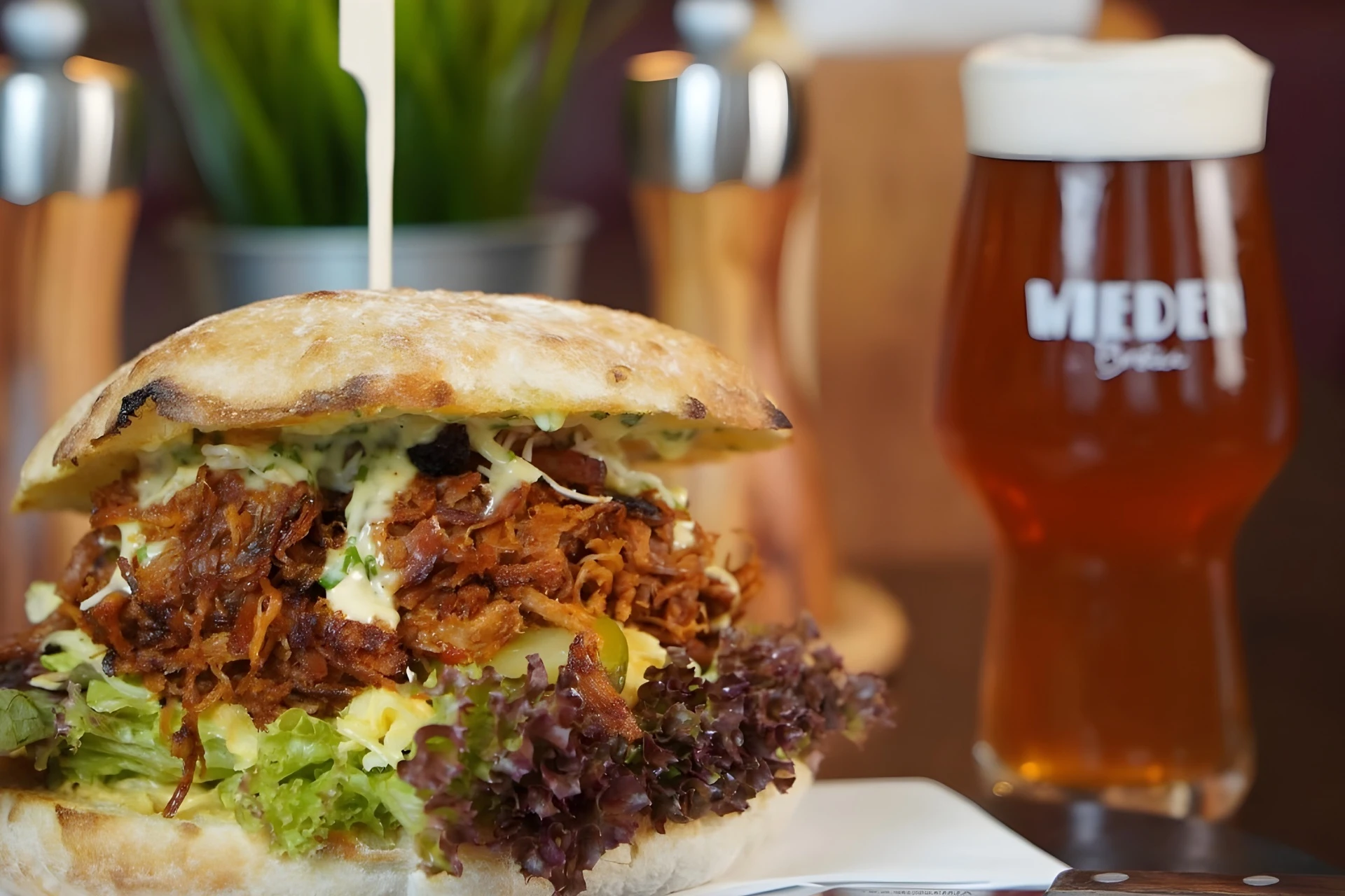 Ein Pulled-Pork-Sandwich mit Salat und Soße liegt auf einem Teller neben einem Glas bernsteinfarbenem Bier; beides steht auf einem Holztisch mit verschwommenem Hintergrund.