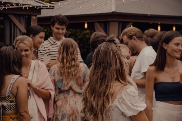 Eine Gruppe von Menschen sitzt bei einem zwanglosen Treffen im Freien zusammen, plaudert und lächelt. Warme Beleuchtung und Lichterketten schaffen eine gemütliche Atmosphäre. Die meisten tragen Sommerkleidung.