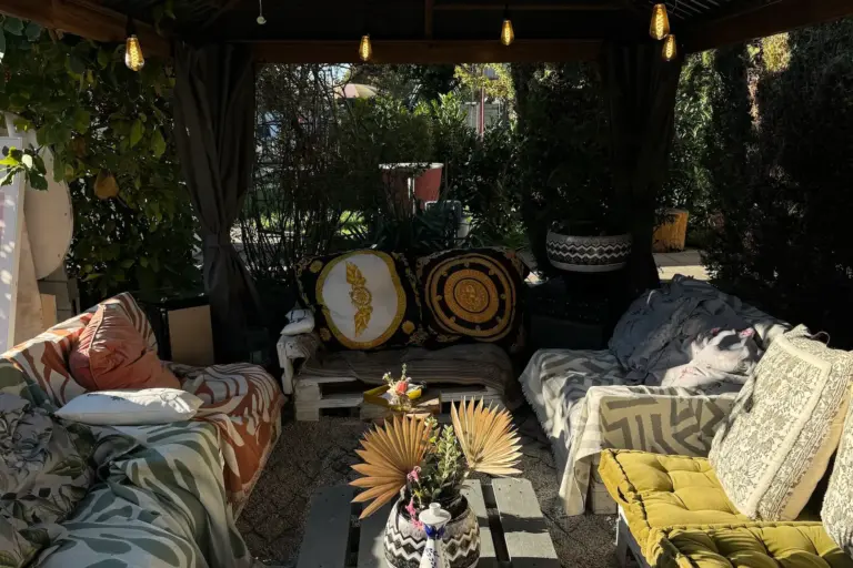Eine gemütliche Sitzecke im Freien mit gepolsterten Sofas, gemusterten Kissen, einem niedrigen Tisch, der mit Vasen und Palmenblättern dekoriert ist, und Lichterketten, die von einer hölzernen Pergola herabhängen, umgeben von Grünpflanzen.