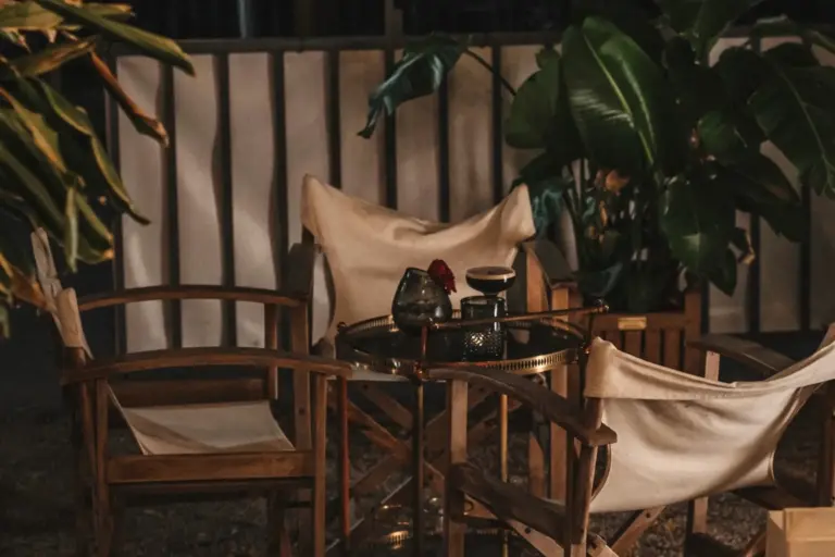Ein gemütlicher nächtlicher Sitzplatz im Freien mit Holzstühlen, einem runden Tisch mit einer Kerze und einer Blumenvase, umgeben von üppigen Grünpflanzen und sanfter Beleuchtung.