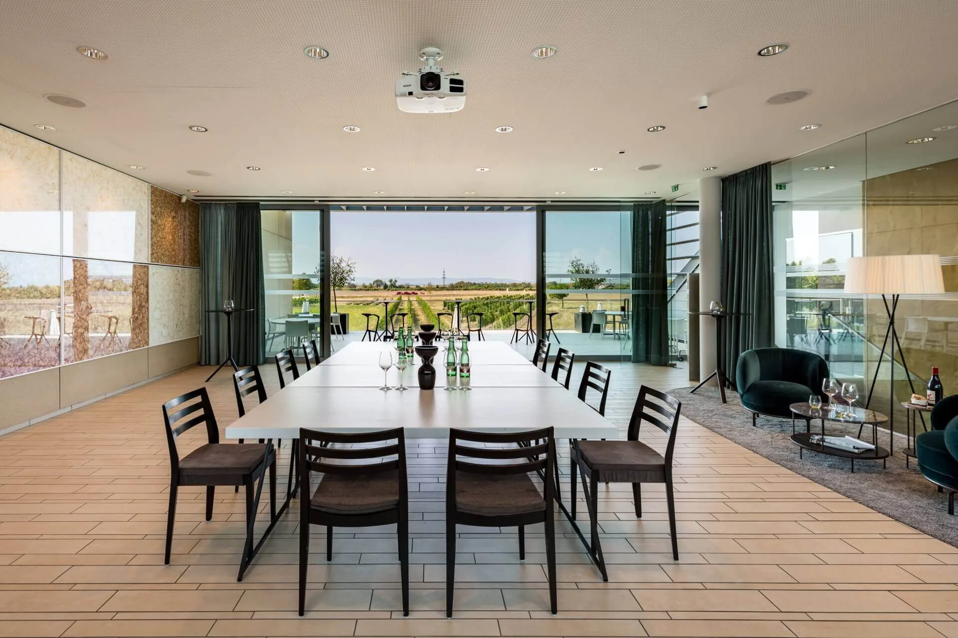 Ein moderner Besprechungsraum mit einem langen Tisch, der von Stühlen, Weingläsern und Flaschen umgeben ist, großen Fenstern, die den Blick nach draußen freigeben, und Loungesitzen im Hintergrund.