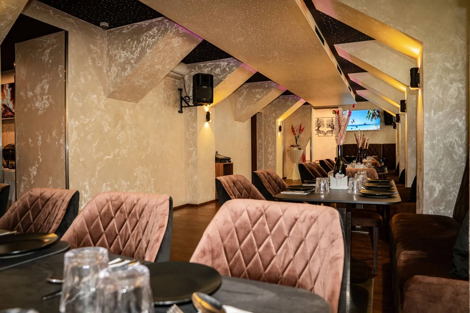 Die elegante Einrichtung des Restaurants mit plüschigen, gesteppten braunen Stühlen, ordentlich eingedeckten Tischen mit schwarzen Tellern und Gläsern, strukturierten beigen Wänden, einer einzigartigen schrägen Decke und moderner Beleuchtung schafft eine warme, einladende Atmosphäre.