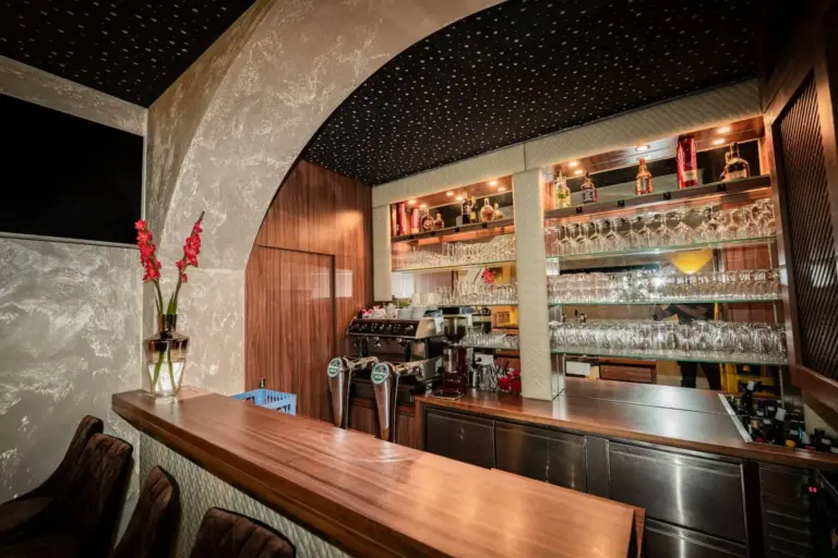 Eine stilvolle Bar mit einem Holztresen, Regalen mit Gläsern und Flaschen, einer Kaffeemaschine und Bierzapfanlagen. Die Decke ist schwarz mit kleinen weißen Punkten, und auf dem Tresen steht eine Vase mit roten Blumen.