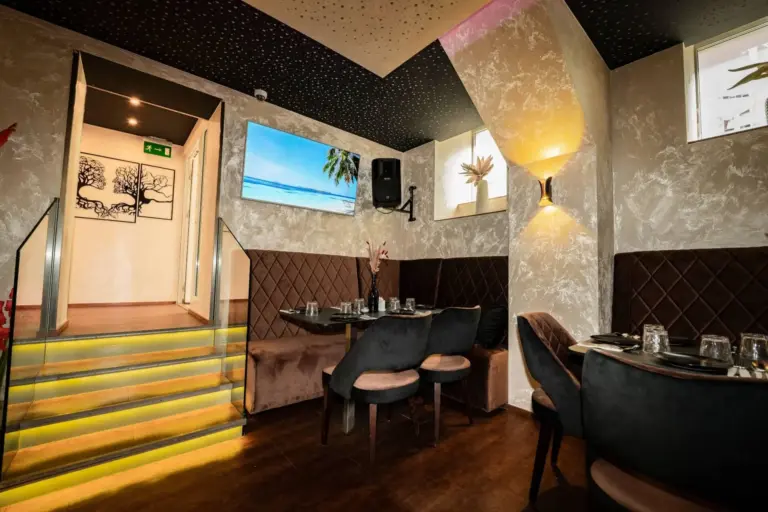 Ein modernes Restaurant mit gepolsterten Stühlen und dunklen Tischen, warmer Beleuchtung, einer gelb beleuchteten Treppe, beigefarbenen Wänden mit Textur und einer Wandmalerei mit einer Strandszene und Palmenblättern.