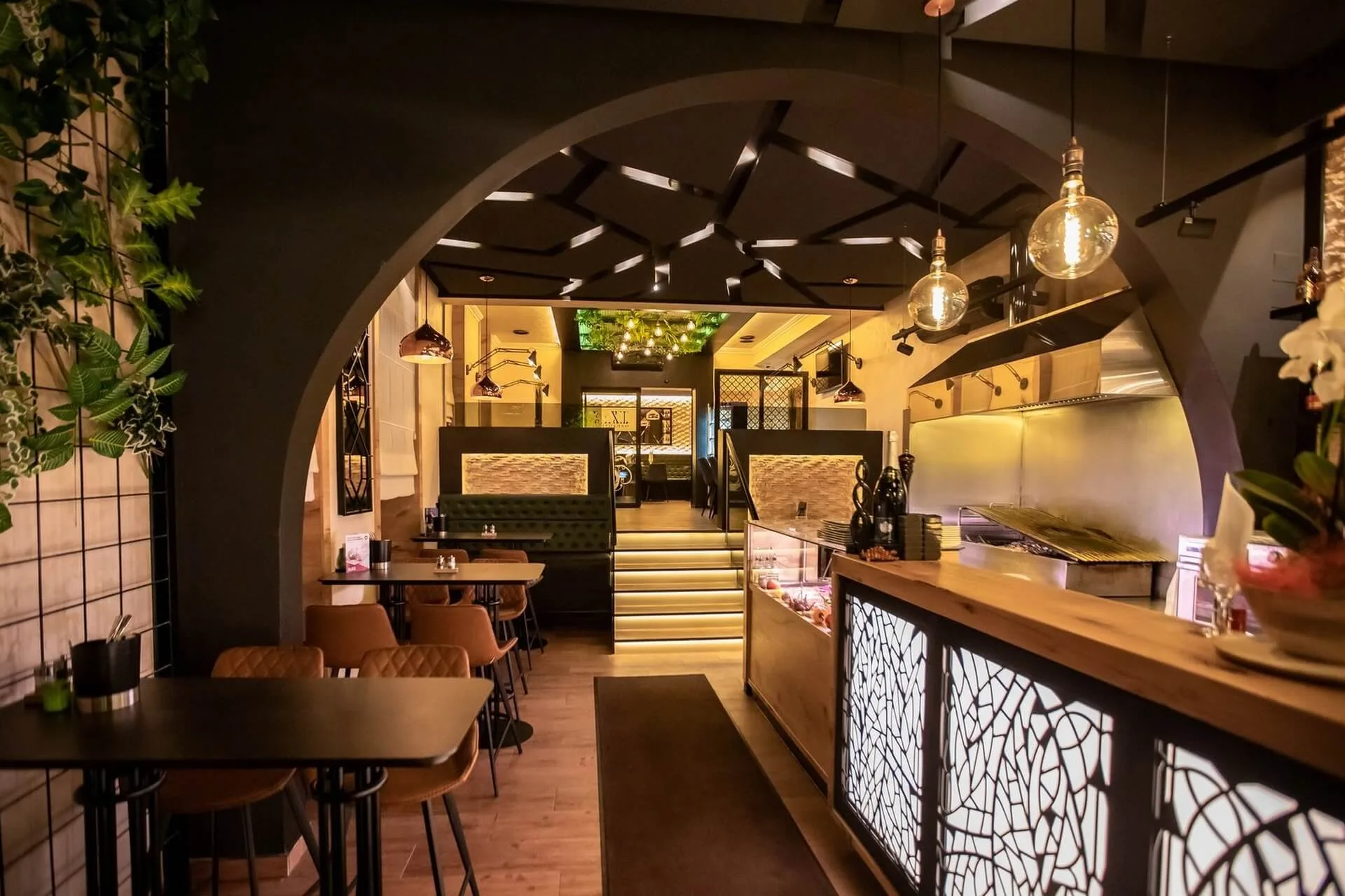 Modernes Restaurantinterieur mit warmer Beleuchtung, Holztischen und -stühlen, dekorativen Hängelampen, Grünpflanzen an den Wänden und einer bogenförmigen Öffnung, die zu einem Essbereich mit Ständen und Stufen im Hintergrund führt.