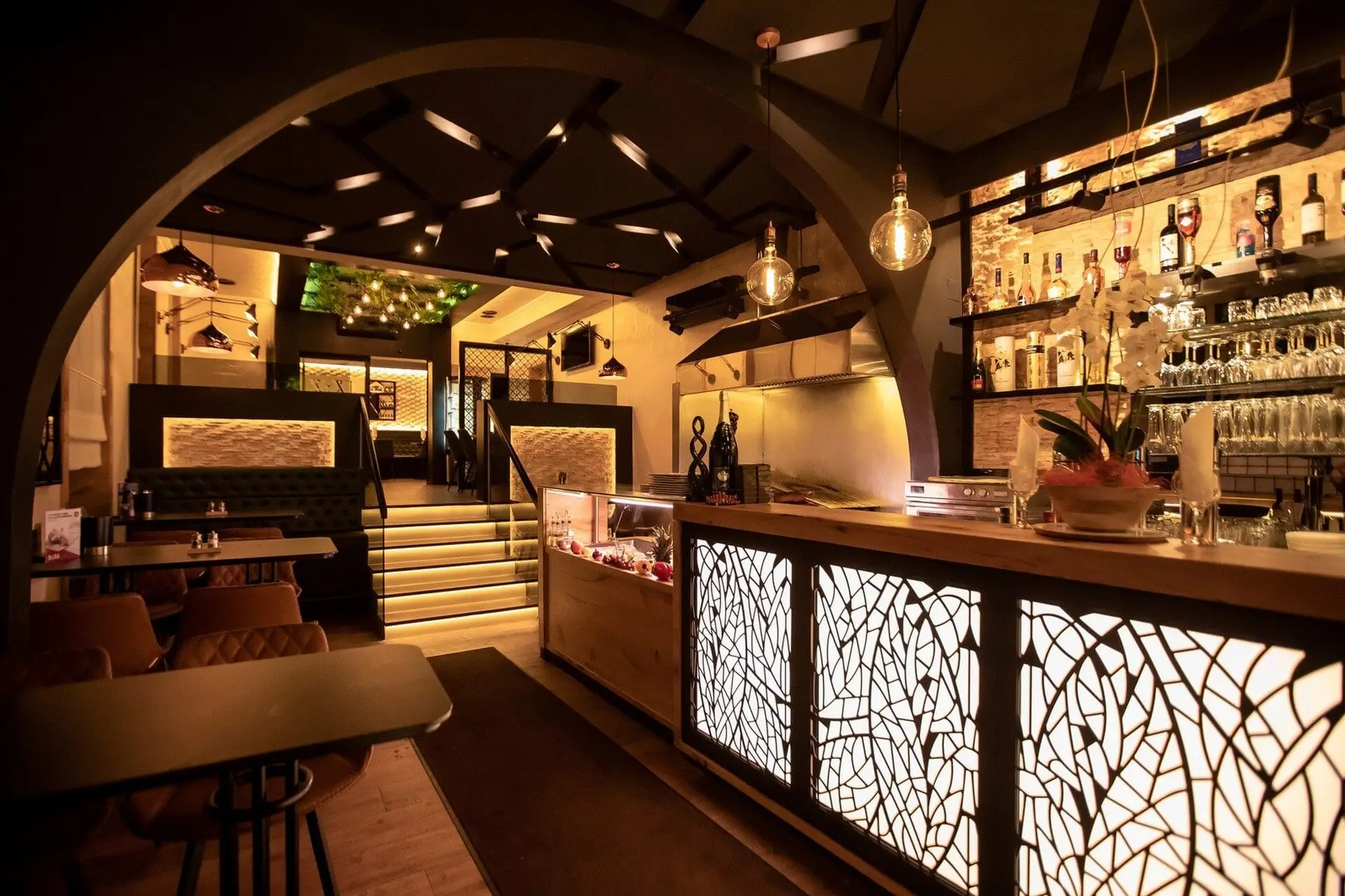 Das moderne, warm beleuchtete Restaurant verfügt über eine dekorative Beleuchtung, gemusterte Paneele, eine Bar mit Regalen mit Flaschen und Gläsern, Tische, gepolsterte Sitzgelegenheiten und eine Treppe, die zu einem oberen Essbereich führt.