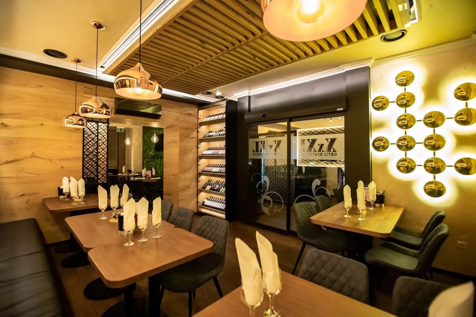 Ein modernes Restaurant mit Holztischen, dunklen gepolsterten Stühlen und Weingläsern mit gefalteten Servietten. Die Weinflaschen sind in Regalen ausgestellt, und die warme Beleuchtung schafft ein gemütliches Ambiente.