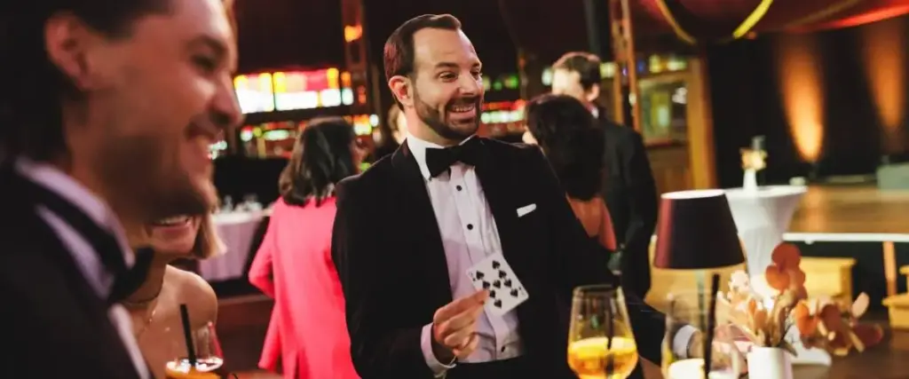 Bei einem Junggesellenabschied in Wien führt ein Mann im Smoking zwei lächelnden Gästen einen Kartentrick vor. Er hält eine Spielkarte hoch, während andere Gäste und elegante Dekorationen den Hintergrund füllen.