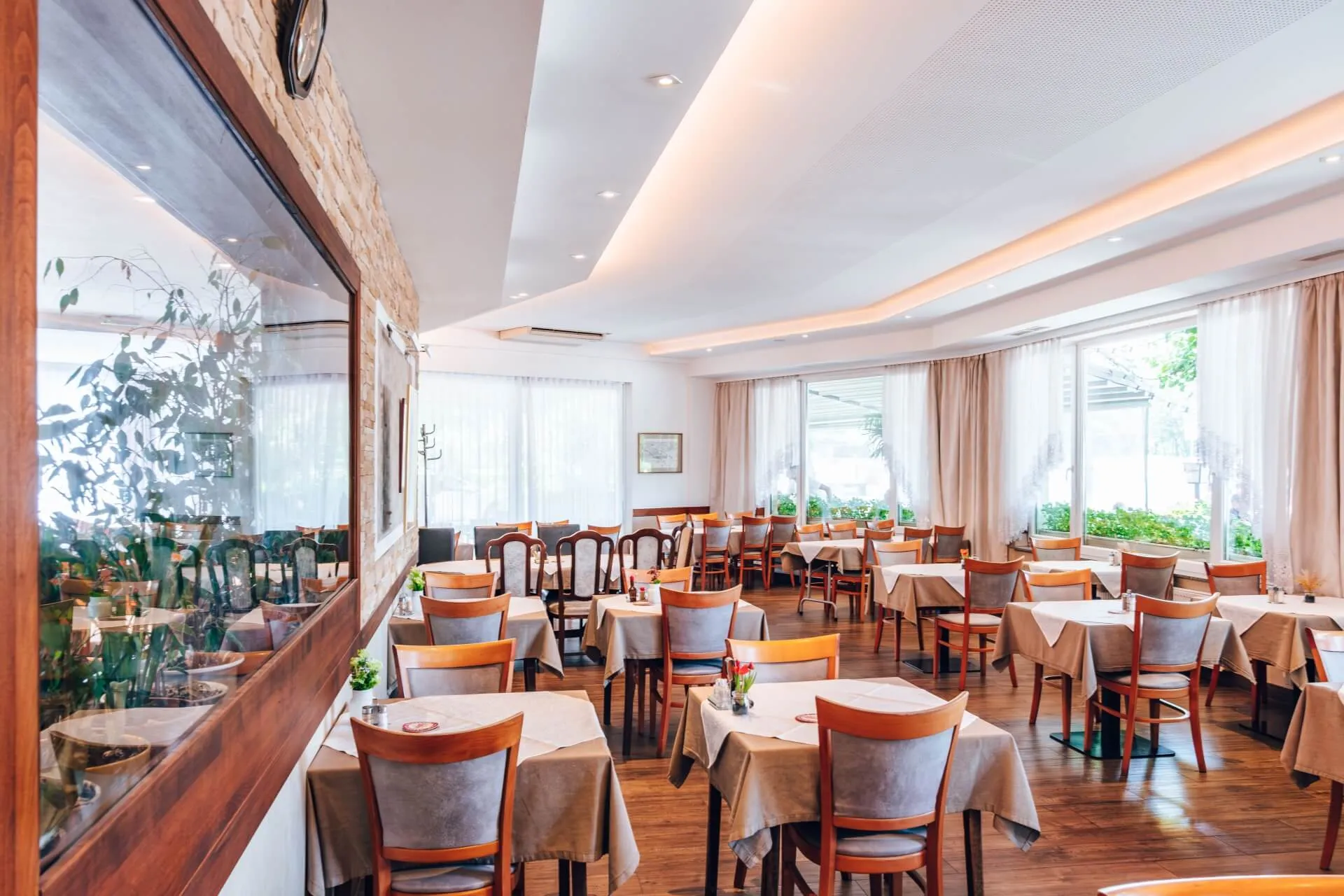 Helles, elegantes Restaurant mit Holzböden, ordentlich gedeckten Tischen mit beigen Tüchern und Holzstühlen. Große Fenster lassen natürliches Licht herein, und der Raum wirkt sauber und einladend.