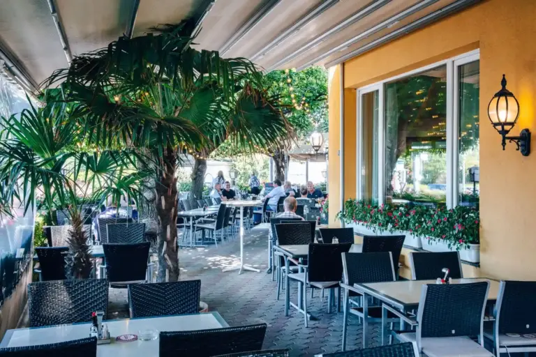 Außenterrasse eines Restaurants mit schwarzen Korbstühlen und weißen Tischen, beschattet von Palmen und einem Sonnensegel. Im Hintergrund sitzen ein paar Leute, und blühende Pflanzen schmücken das Fensterbrett des gelben Gebäudes.