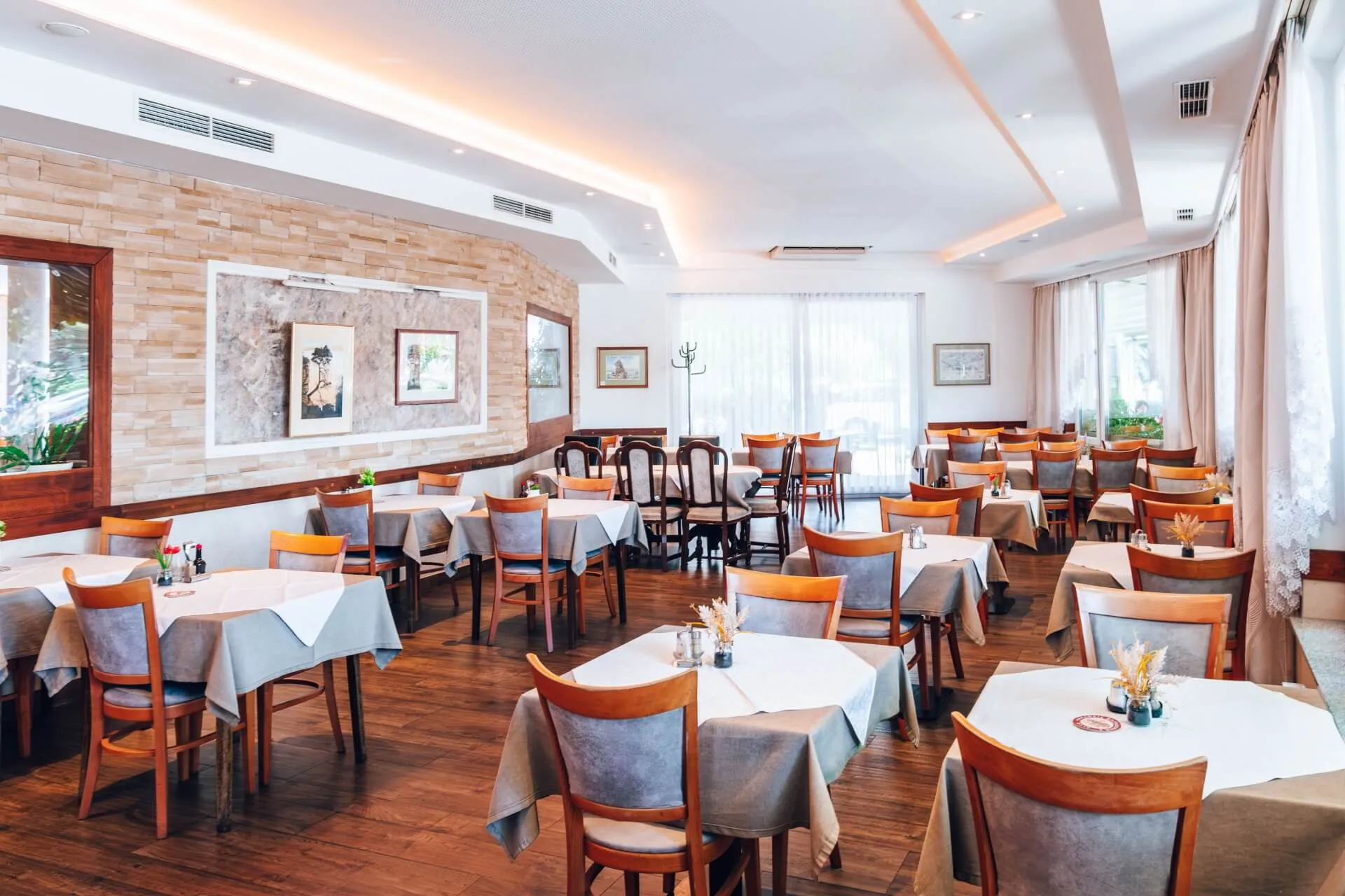 Ein helles, geräumiges Restaurant mit Holzböden, ordentlich aufgestellten Tischen mit weißen und grauen Tischdecken, Holzstühlen, Wandkunst und großen Fenstern, die viel Tageslicht hereinlassen.