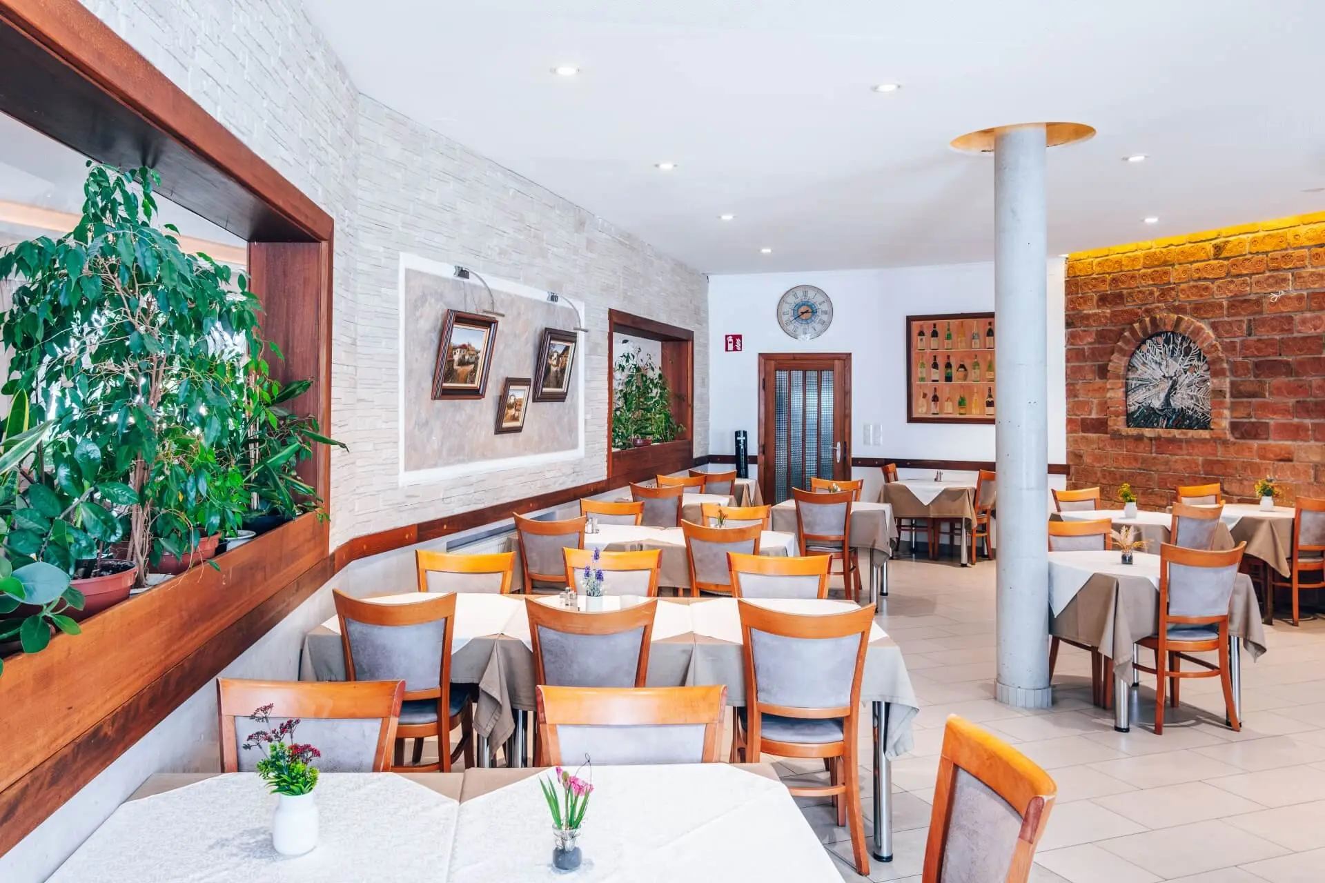 Ein heller, moderner Restaurantbereich mit Holzstühlen und Tischen, die mit weißen Tüchern bedeckt sind, dekoriert mit kleinen Topfpflanzen, Wandkunst und Grünpflanzen, mit natürlichem Licht.