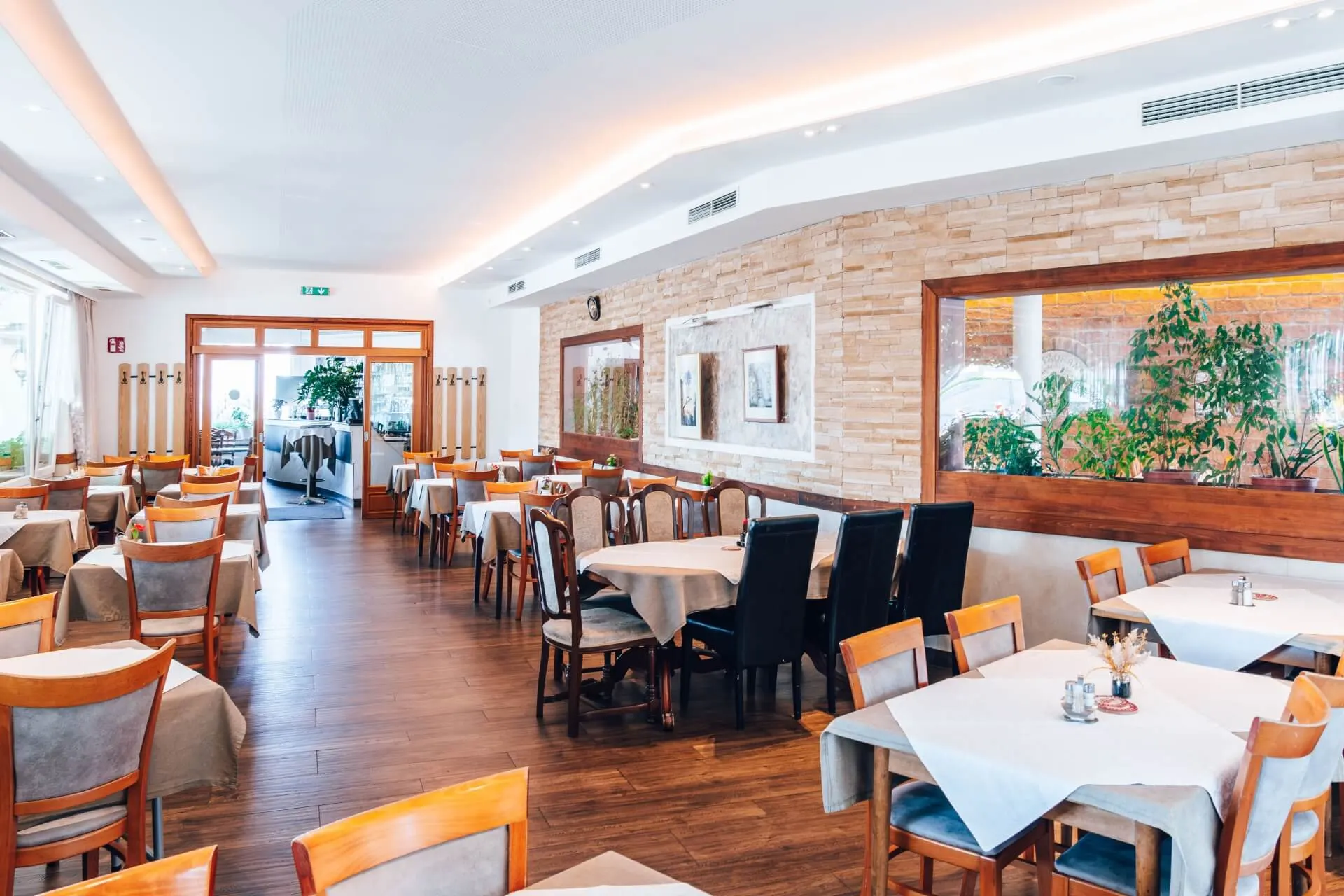 Ein helles, modernes Restaurant mit Holzböden, ordentlich aufgestellten Tischen mit weißen Tischdecken, gepolsterten Stühlen, großen Fenstern und dekorativen Pflanzen an den Wänden.