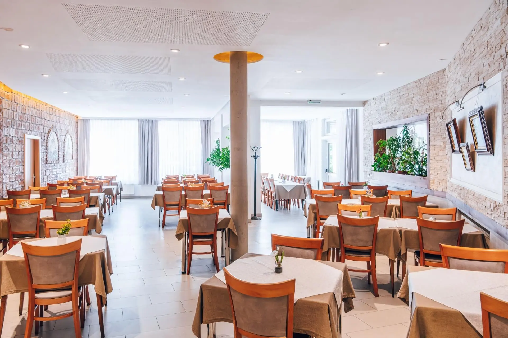 Ein heller, moderner Restaurantbereich mit ordentlich aufgestellten Tischen und Holzstühlen, beigen Tischdecken, großen Fenstern mit Vorhängen, Steinwänden, Pflanzen und zeitgenössischem Dekor.