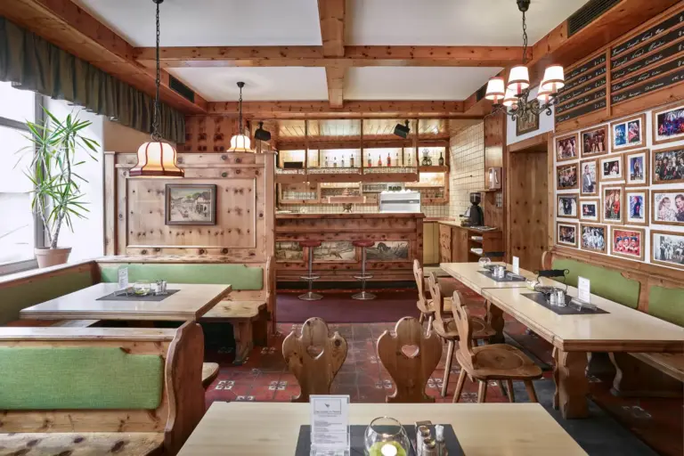 Ein gemütliches, holzgetäfeltes Restaurant mit grün gepolsterten Bänken, Holztischen und -stühlen, Wandfotos, Hängelampen und einem Barbereich im Hintergrund. Durch ein großes Fenster auf der linken Seite fällt natürliches Licht ein.