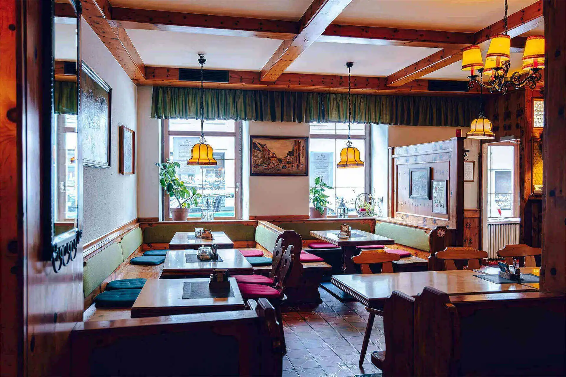 Ein gemütliches, rustikales Restaurant mit Holzbalken, Tischen und Bänken. Das Sonnenlicht strömt durch große Fenster, und darüber hängen gelbe Hängelampen. Rote und grüne Kissen säumen die Sitzbereiche.