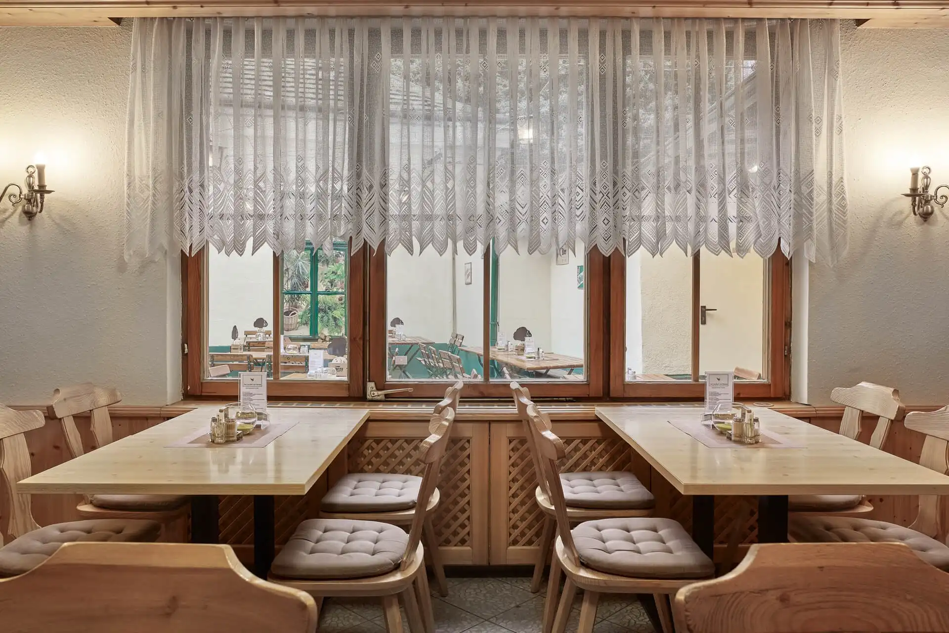 Ein gemütliches Restaurant mit Holztischen und gepolsterten Stühlen, Spitzenvorhängen an den Fenstern und sanfter Beleuchtung. Die Fenster gehen auf einen Außenbereich mit weiteren Tischen und Grünpflanzen hinaus.