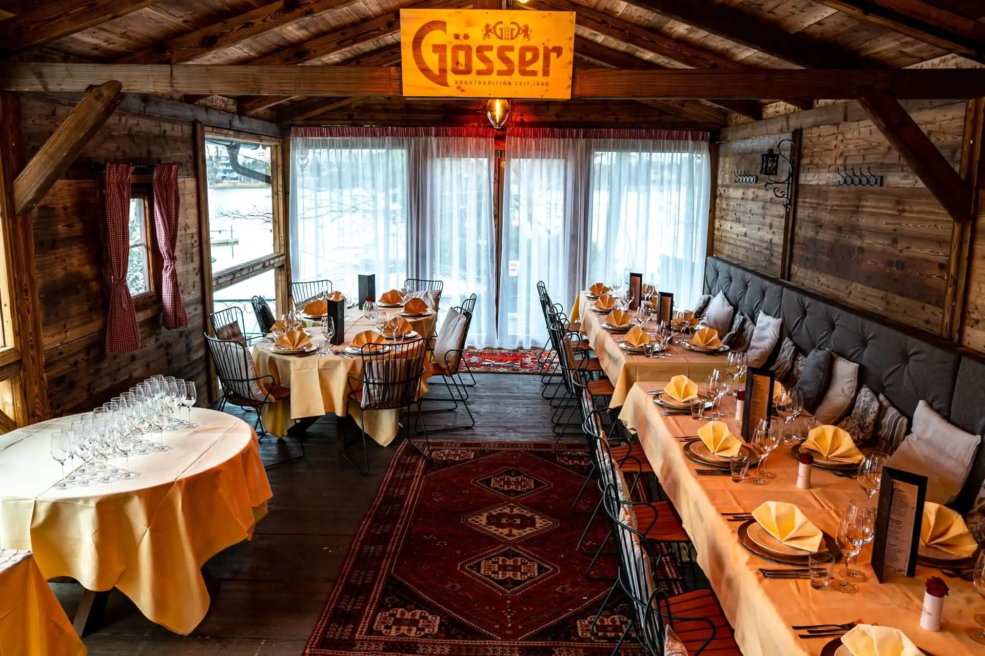 Ein gemütliches, rustikales Restaurant mit hölzernen Wänden und Decken, das mit gelben Servietten und Tischtüchern ausgestattet ist. Große Fenster an der Rückseite lassen natürliches Licht herein, und darüber hängt ein Gösser-Schild. Rote Teppiche bedecken den Holzboden.