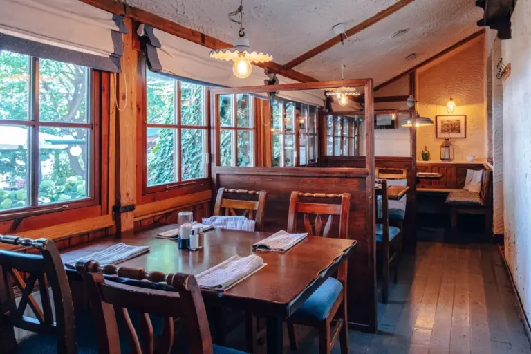 Gemütliches Restaurant mit Holztischen und -stühlen, ordentlich platzierten Servietten, großen Fenstern, die natürliches Licht hereinlassen, und rustikaler Einrichtung mit Hängelampen und Holzverkleidung.